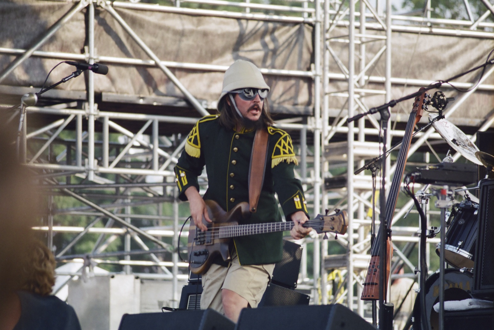 A bassist in a pith helmet, goggle sunglasses and a green military jacket with gold epaulettes plays on a scaffolded festival stage.